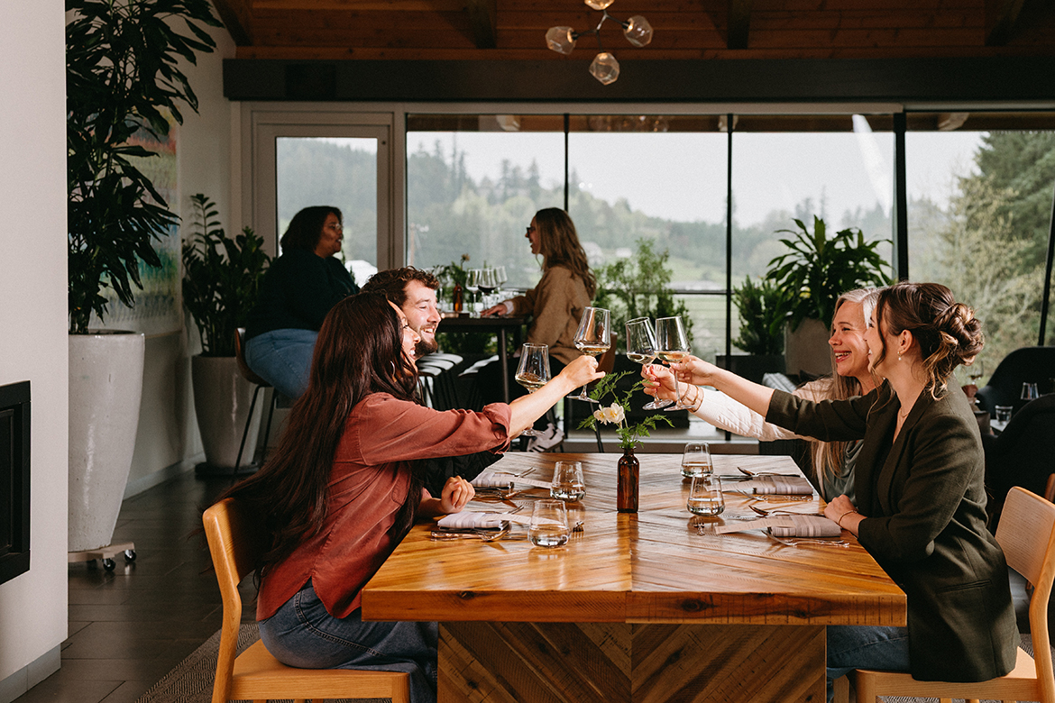 Four people sitting in the Rex Hill Tasting Room, raising there glasses to cheers, while other visitors are seated in the background.