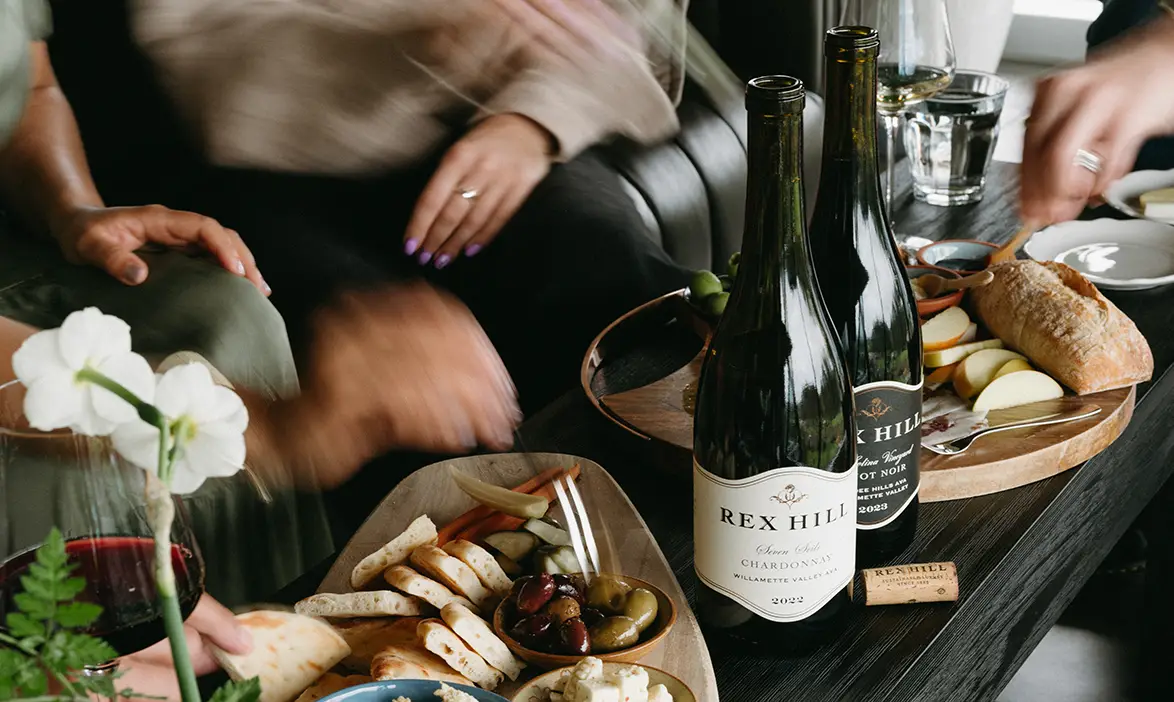 Rex Hill wines place on a coffee table with charcuterie board. Three people are sat around the table enjoying their wine and food.